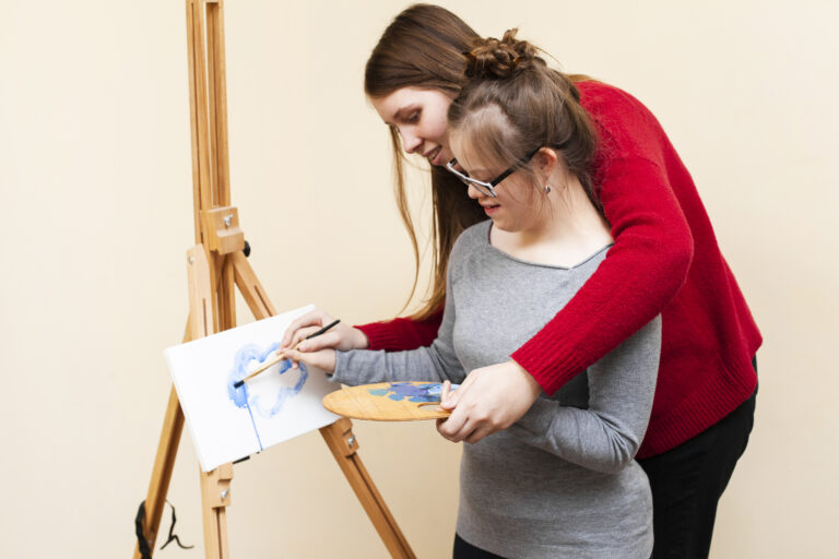 Side View Woman Helping Girl With Down Syndrome Paint 1 768x512